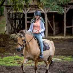 Dziewczyna w kasku jeździ na koniu rasy fiording na ujeżdżalni; w tle drewniana stajnia i drzewa.