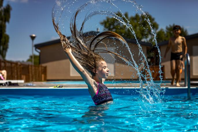 Dziewczynka w stroju kąpielowym odrzuca mokre włosy w basenie podczas słonecznego dnia, w tle budynek kolonijny i drugi chłopiec.
