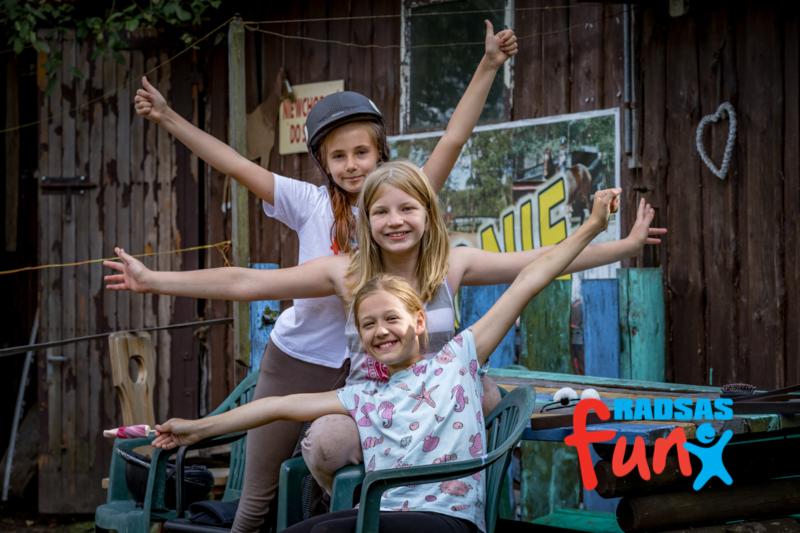 Trzy uśmiechnięte dziewczynki pozują z rozłożonymi ramionami na tle drewnianego budynku z napisem i dekoracją w kształcie serca, jedna z nich ma kask na głowie.