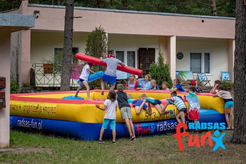 Dzieci w trakcie zabawy na dmuchanym ringu z piankowymi pałkami, w tle budynek z oknami i drzewa.