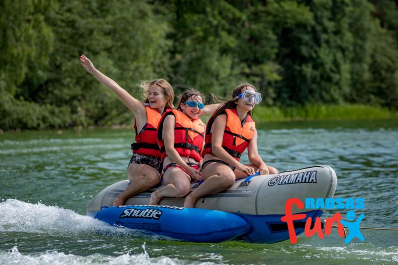 Trzy nastolatki w kamizelkach ratunkowych siedzą na dmuchanym bananie ciągniętym po jeziorze, jedna z dziewcząt macha ręką, w tle zielone drzewa.