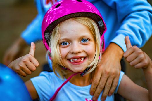 Uśmiechnięta dziewczynka w różowym kasku, z widocznymi brakami w uzębieniu, pokazuje kciuki w górę, wspierana ramieniem dorosłego.