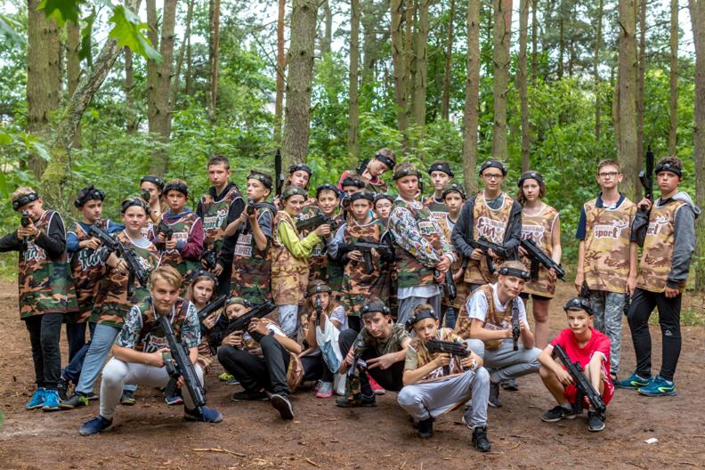 Grupa młodzieży w lesie ubrana w kamuflażowe stroje, trzymająca pistolety do laserowego paintballa, pozująca do zdjęcia.