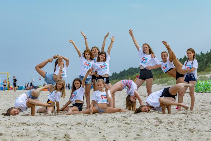 Grupa dziewcząt w białych koszulkach z logo na plaży, prezentujących figury gimnastyczne i taneczne, z drzewami w tle.
