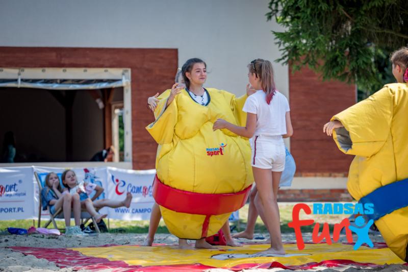 Dziewczyna w żółtym stroju sumo z czerwonym pasem rozmawia z inną dziewczyną na piasku podczas zajęć rekreacyjnych.