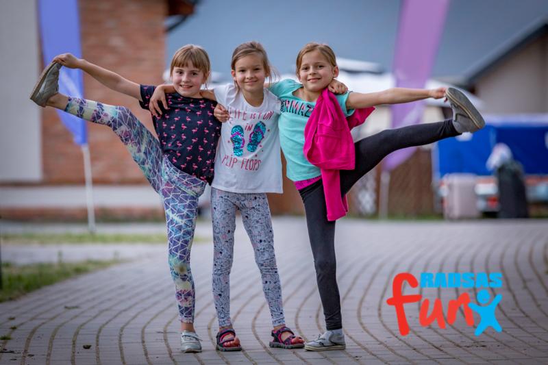 Trzy uśmiechnięte dziewczynki w różnokolorowych strojach, stojące ramię w ramię na brukowanej drodze, dwie z nich z wyciągniętymi nogami w bok, w tle budynek i flaga.