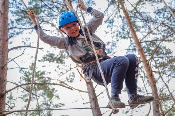 Uśmiechnięty chłopiec w niebieskim kasku, wiszący na linach w parku linowym, z widocznym uprzężą i drzewami w tle.