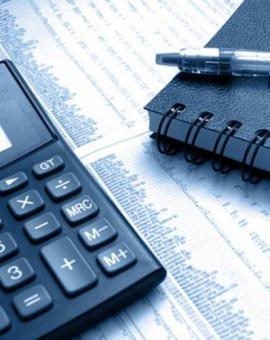 Kalkulator, notatnik i długopis leżące na arkuszu z danymi liczbowymi w odcieniach niebieskiego. Widoczne przyciski kalkulatora i faktura papieru.