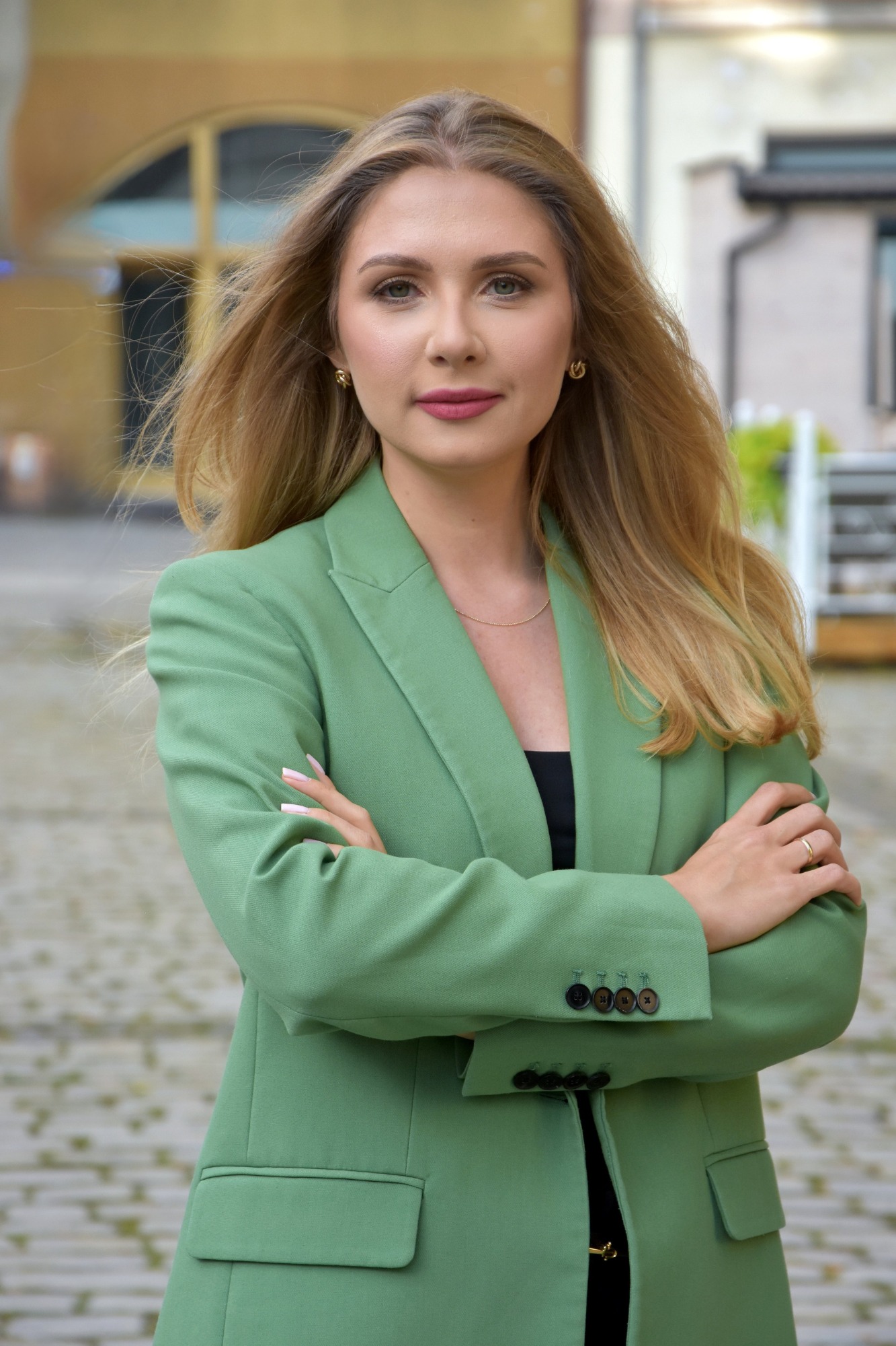 Portret kobiety w zielonej marynarce z założonymi rękami na tle brukowanej ulicy i budynku