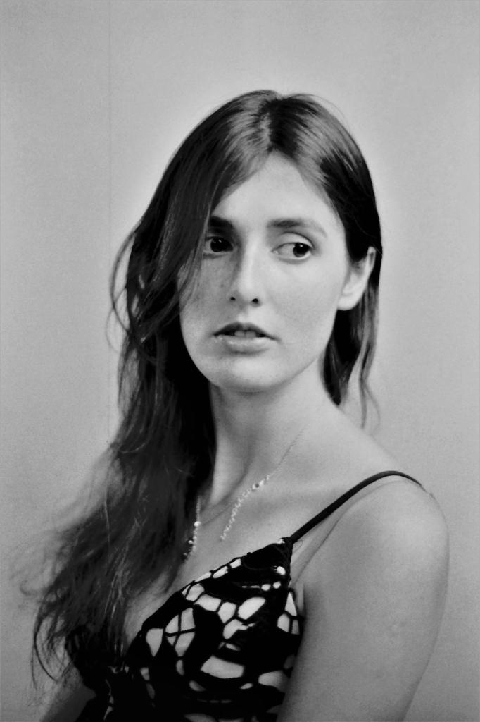 Portret kobiety z długimi, ciemnymi włosami, patrzącej w bok, w czarno-białej sukience na ramiączkach z geometrycznym wzorem i delikatnym naszyjnikiem, na neutralnym tle.