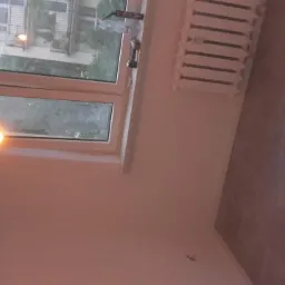 Pokój w Katowicach po remoncie: świeżo pomalowane ściany w odcieniach bieli, widoczne nowe panele podłogowe w kolorze szarości, okno z widokiem na zieleń i ozdobna lampa wisząca.