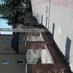 Widok z zewnątrz na dom z nową elewacją i brukowanym chodnikiem z czerwonymi akcentami, widoczny słup energetyczny i drzewa w tle.