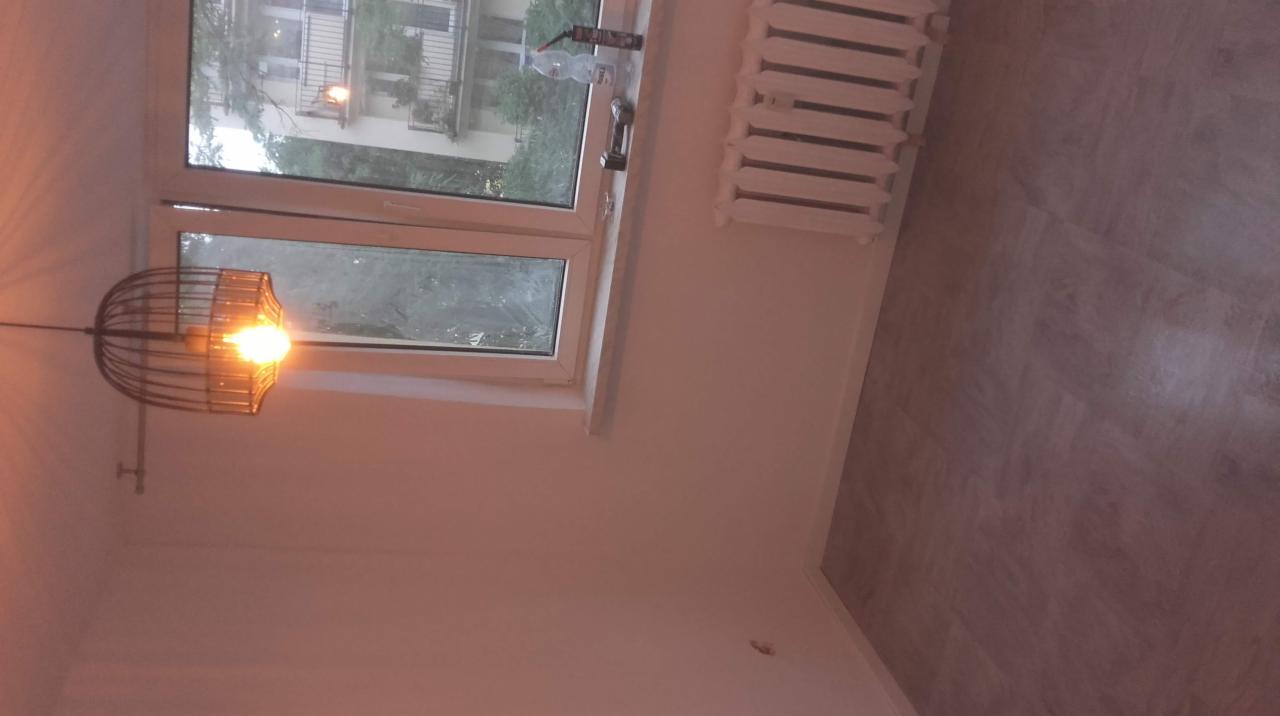 Pokój w Katowicach po remoncie: świeżo pomalowane ściany w odcieniach bieli, widoczne nowe panele podłogowe w kolorze szarości, okno z widokiem na zieleń i ozdobna lampa wisząca.