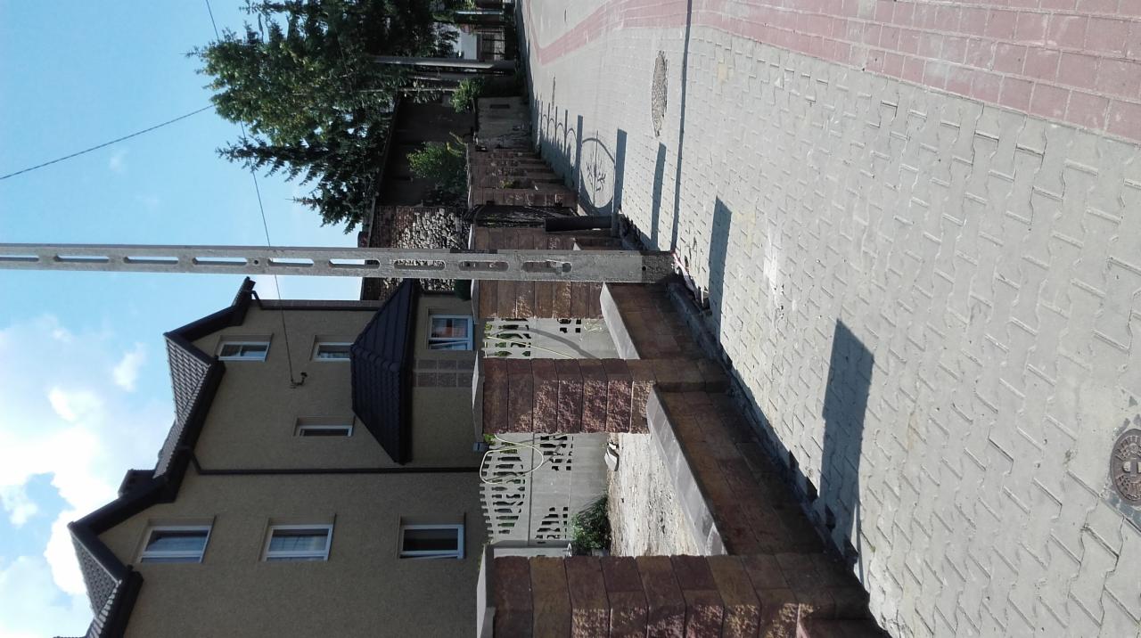 Widok z zewnątrz na dom z nową elewacją i brukowanym chodnikiem z czerwonymi akcentami, widoczny słup energetyczny i drzewa w tle.