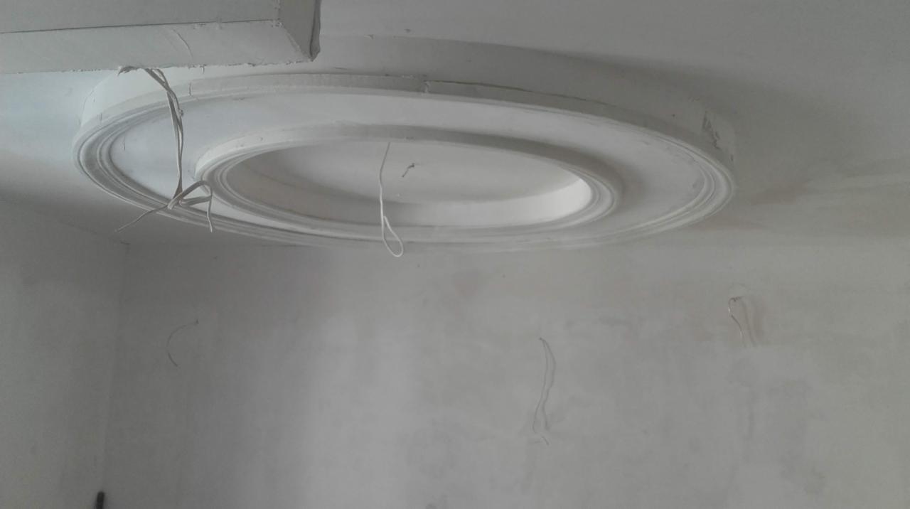 Okrągła, biała dekoracja sufitowa w trakcie montażu, widoczne wystające kable i niedoskonałości na ścianie.