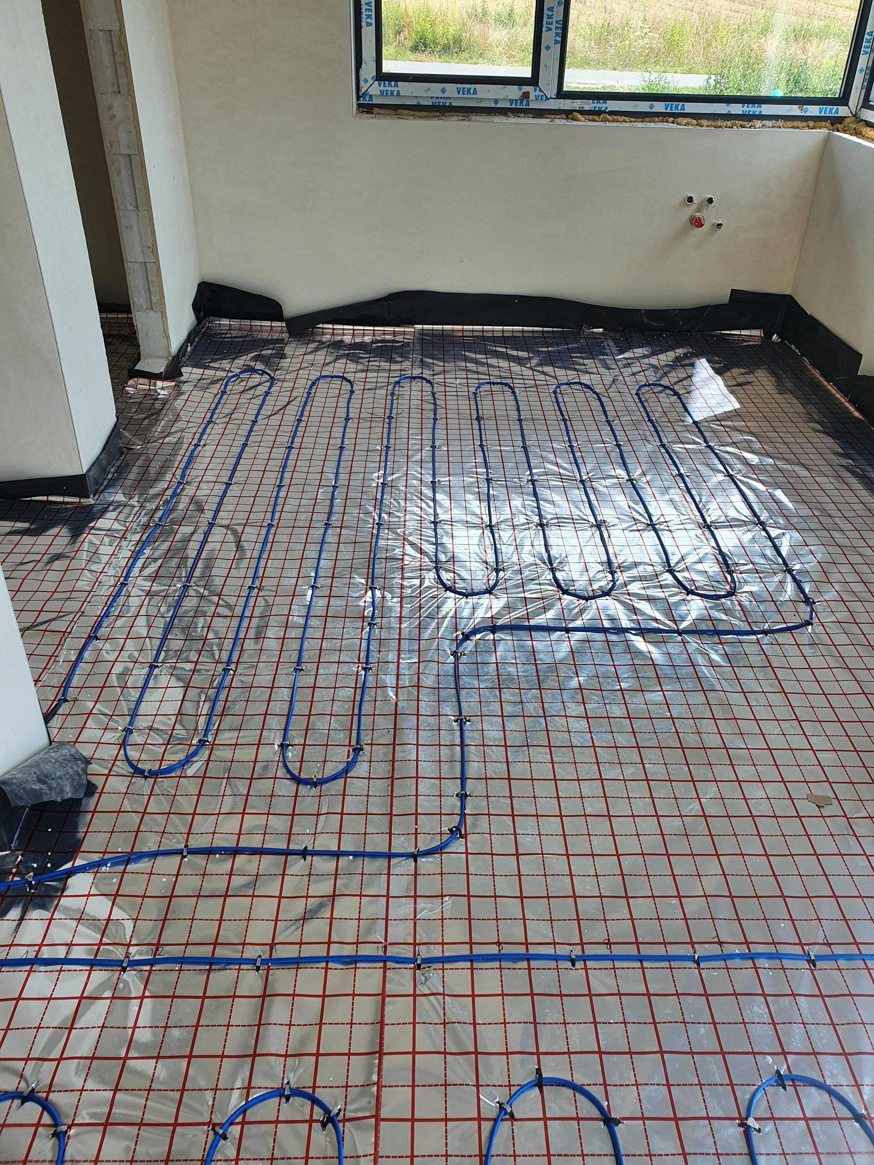 Niebieskie rurki ogrzewania podłogowego ułożone na folii z czerwoną siatką w surowym wnętrzu, widok z góry.