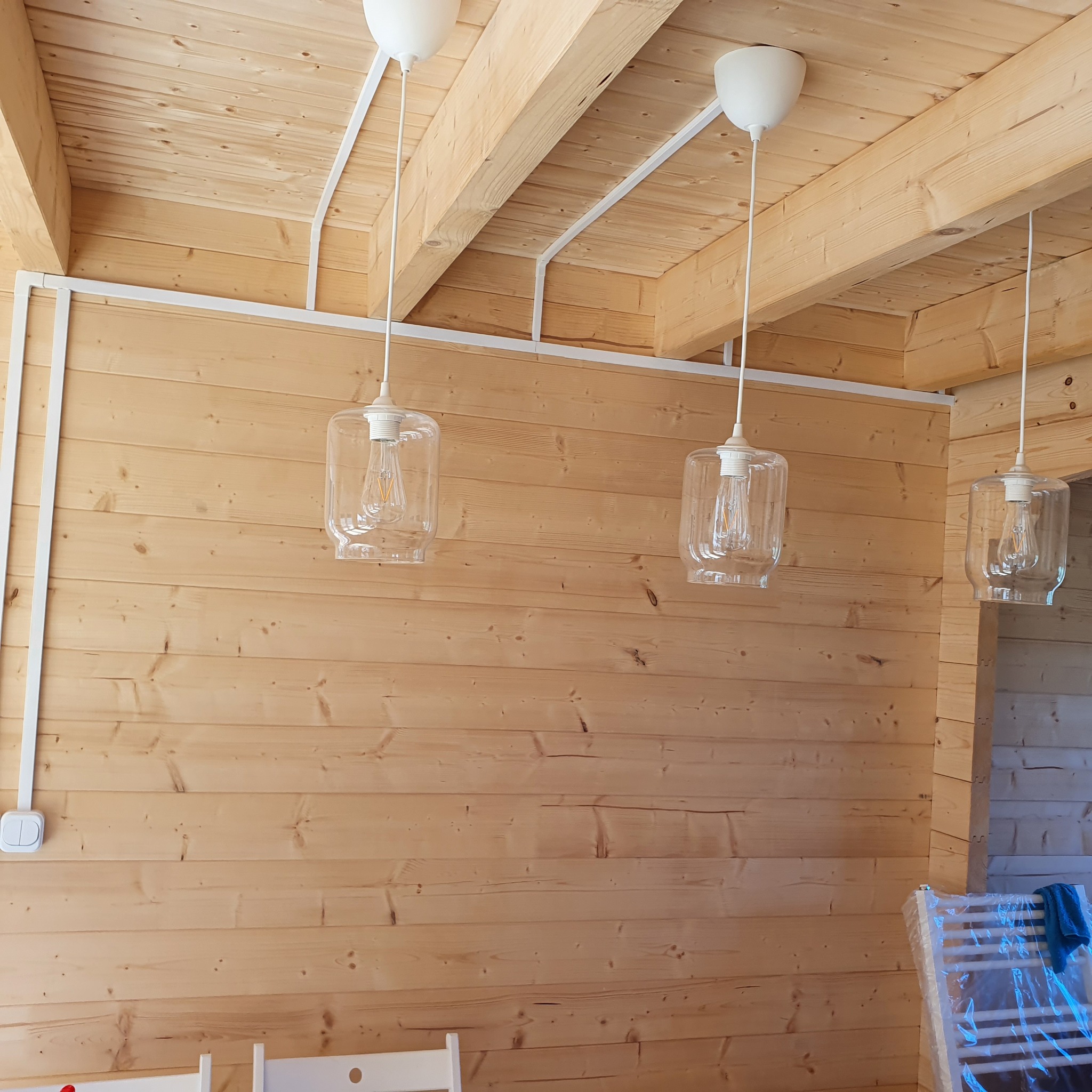 Trzy szklane lampy wiszące z żarówkami Edisona o ciepłej barwie, zamontowane na drewnianym suficie podwieszanym z widocznymi belkami konstrukcyjnymi i białymi listwami kablowymi, w jasnym wnętrzu...