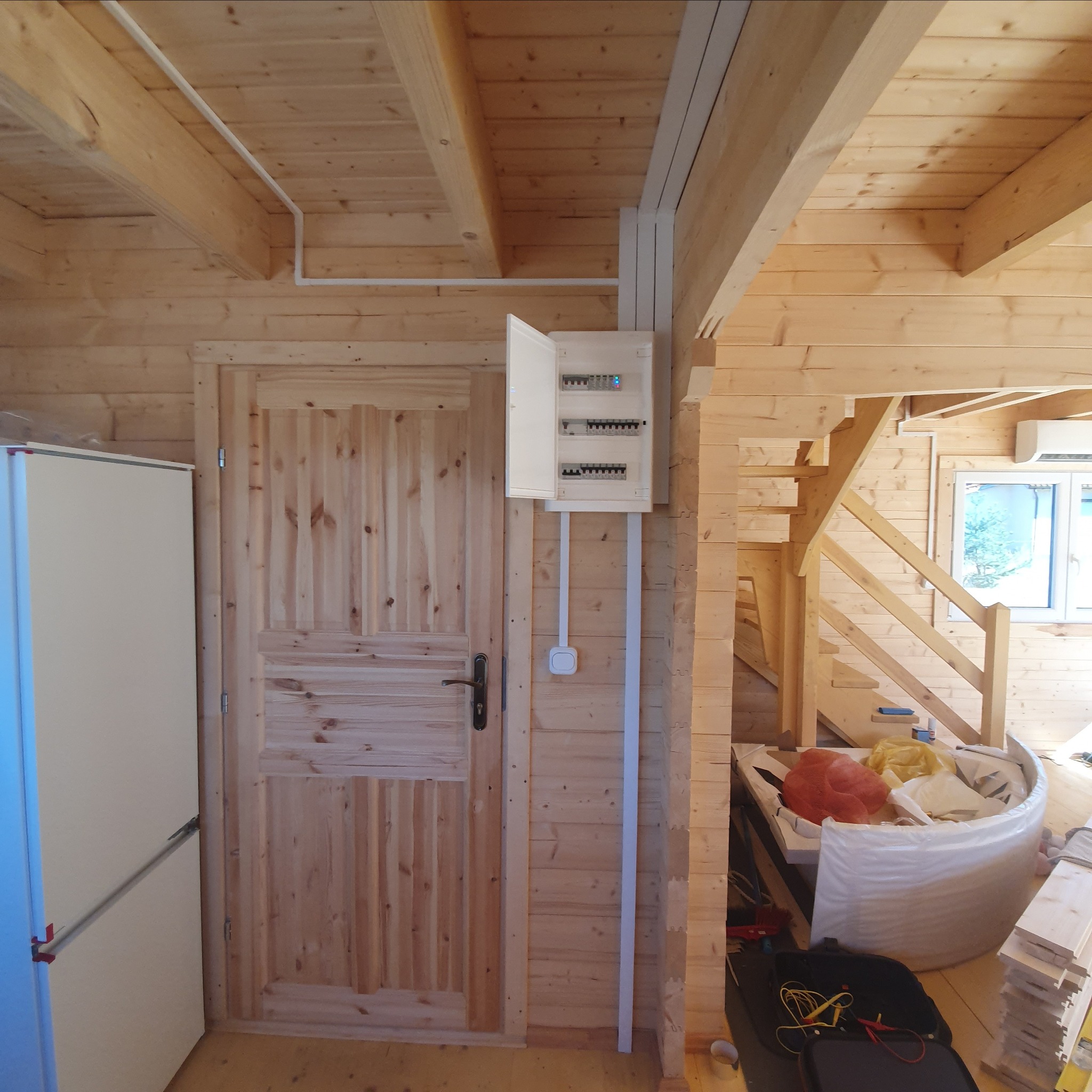 Rozdzielnica elektryczna z otwartymi drzwiczkami zamontowana na ścianie z boazerii w drewnianym domu, widoczne przewody i osprzęt elektryczny, obok drzwi i schody w tle.