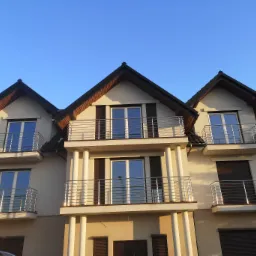 Elewacja trzypiętrowego domu z balkonami zabezpieczonymi metalowymi barierkami ze stali nierdzewnej, widziana z dołu pod kątem w słoneczny dzień.