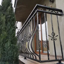 Czarna, metalowa balustrada balkonowa z dekoracyjnymi elementami, widoczna z zewnątrz, z tujami w tle.