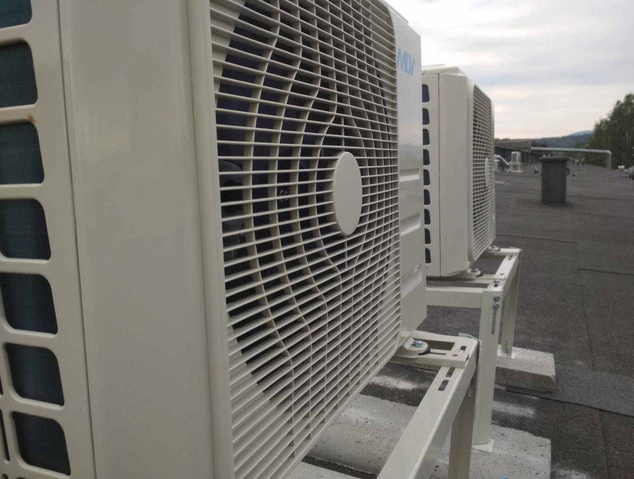 Zewnętrzne jednostki klimatyzacyjne zamontowane na metalowych stelażach na płaskim dachu budynku, widok z boku, pochmurne niebo w tle.