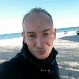 Portret mężczyzny na plaży, z błękitnym niebem i morzem w tle, ubrany w czarną kurtkę.