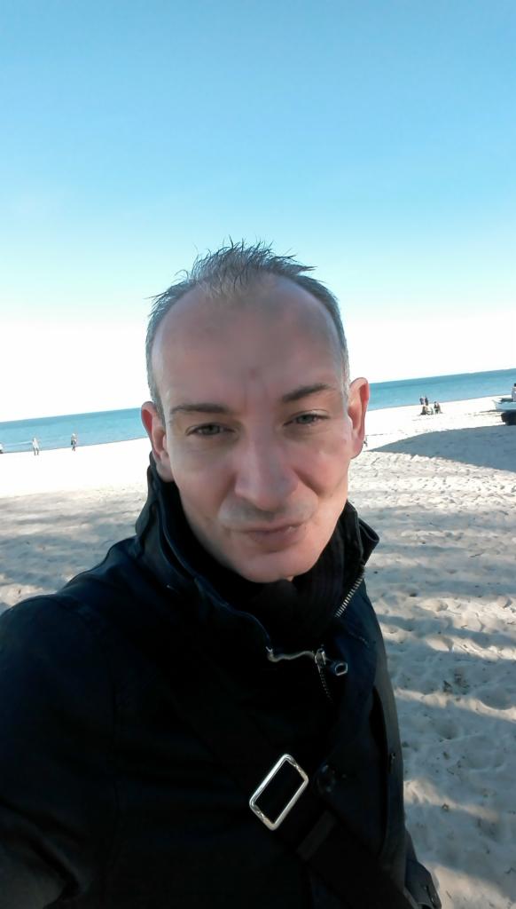 Portret mężczyzny na plaży, z błękitnym niebem i morzem w tle, ubrany w czarną kurtkę.