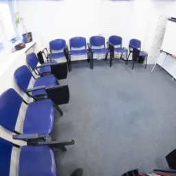 Sala szkoleniowa z niebieskimi krzesłami ustawionymi w okrąg, tablica suchościeralna i okno z żaluzjami w tle, szeroki kąt widzenia.