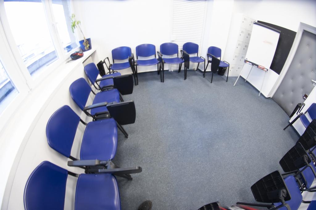 Sala szkoleniowa z niebieskimi krzesłami ustawionymi w okrąg, tablica suchościeralna i okno z żaluzjami w tle, szeroki kąt widzenia.