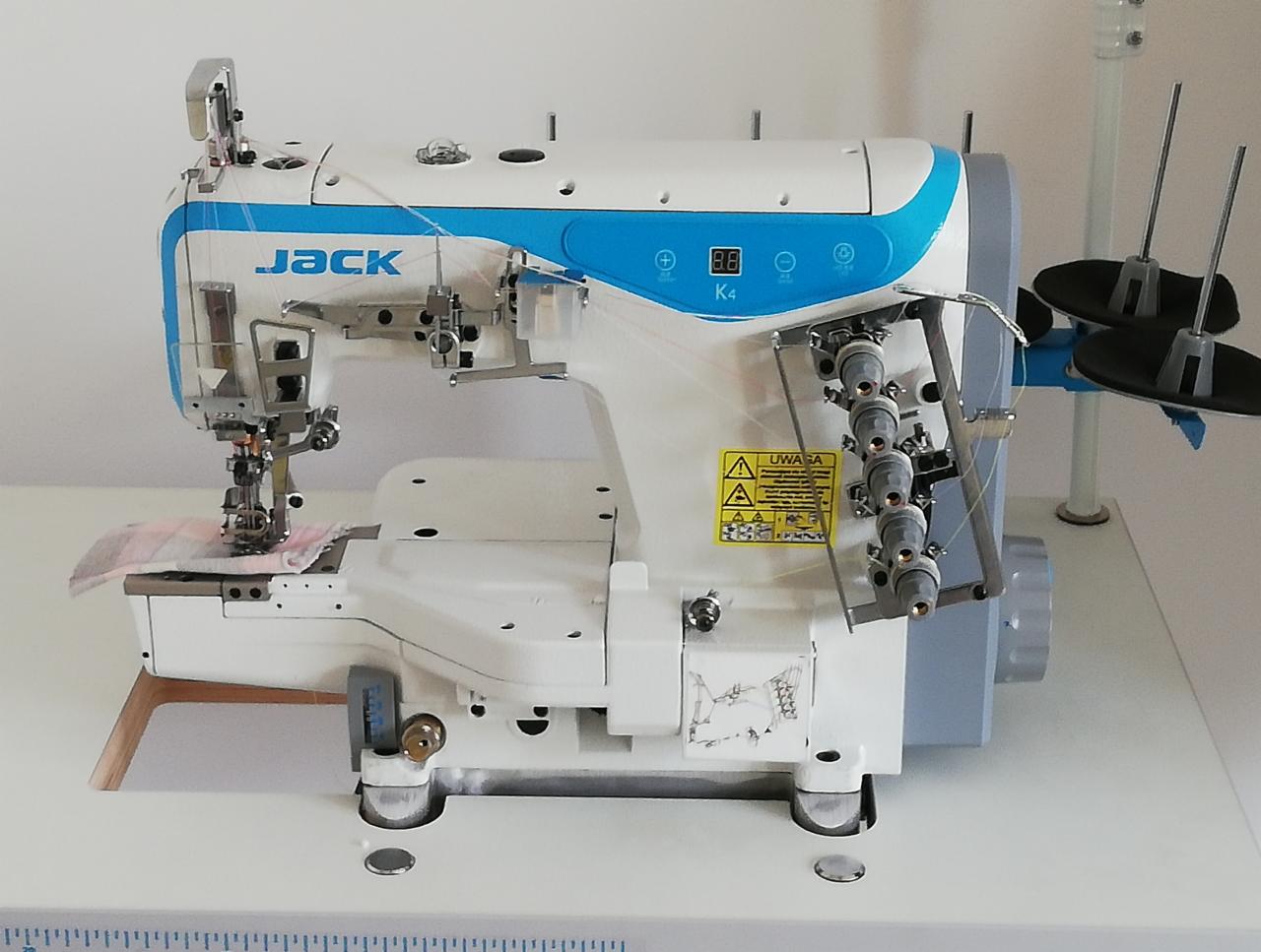 Overlock marki Jack K4 z czterema nićmi podczas szycia wzorzystego materiału, widoczna skala pomiarowa na stole.