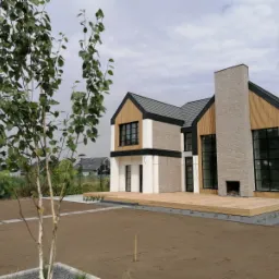 Nowoczesny dom jednorodzinny z drewnianym tarasem, elewacją z cegły i drewna, oraz młodą brzozą na pierwszym planie, w trakcie zagospodarowywania terenu.