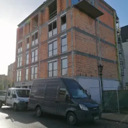 Wielopiętrowy budynek mieszkalny w trakcie budowy z czerwonej cegły, z częściowo zamontowanymi oknami, widoczny z ulicy, z zaparkowanymi samochodami dostawczymi.