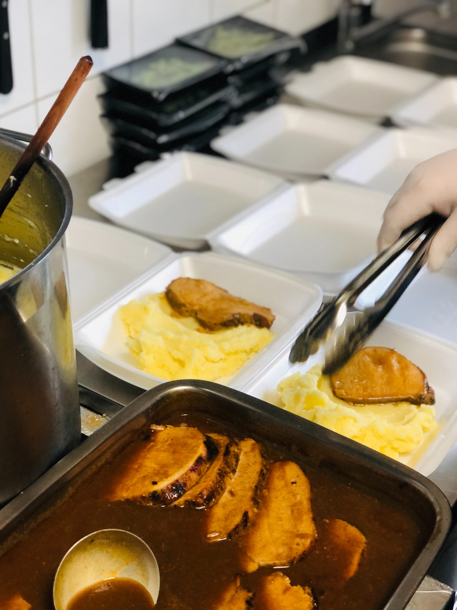 Przygotowywanie porcji mięsa z puree ziemniaczanym do pakowania w białe pudełka, widoczne szczypce w dłoni w rękawiczce, sos w metalowej brytfannie i nabierka.