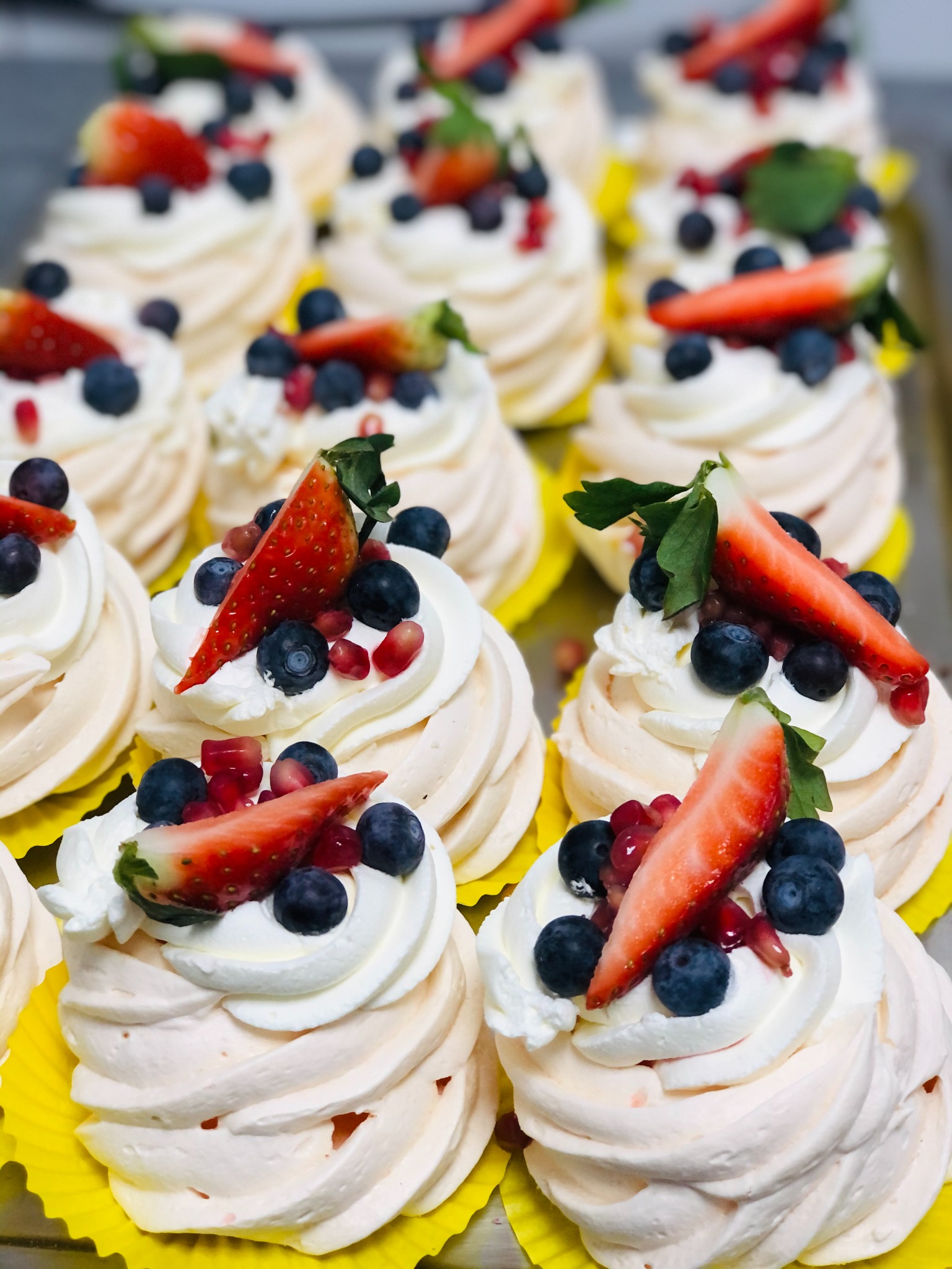 Rząd mini bezowych tortów Pavlova w żółtych papilotkach, udekorowanych bitą śmietaną, borówkami, granatem i truskawkami w kształcie łódeczek z zielonymi listkami.