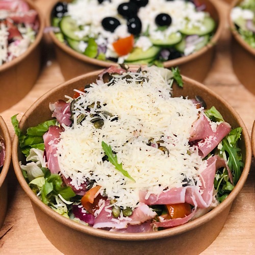 Sałatka z szynką parmeńską, serem i pestkami dyni w papierowej misce, widoczna w rzędzie innych sałatek, oświetlenie naturalne.