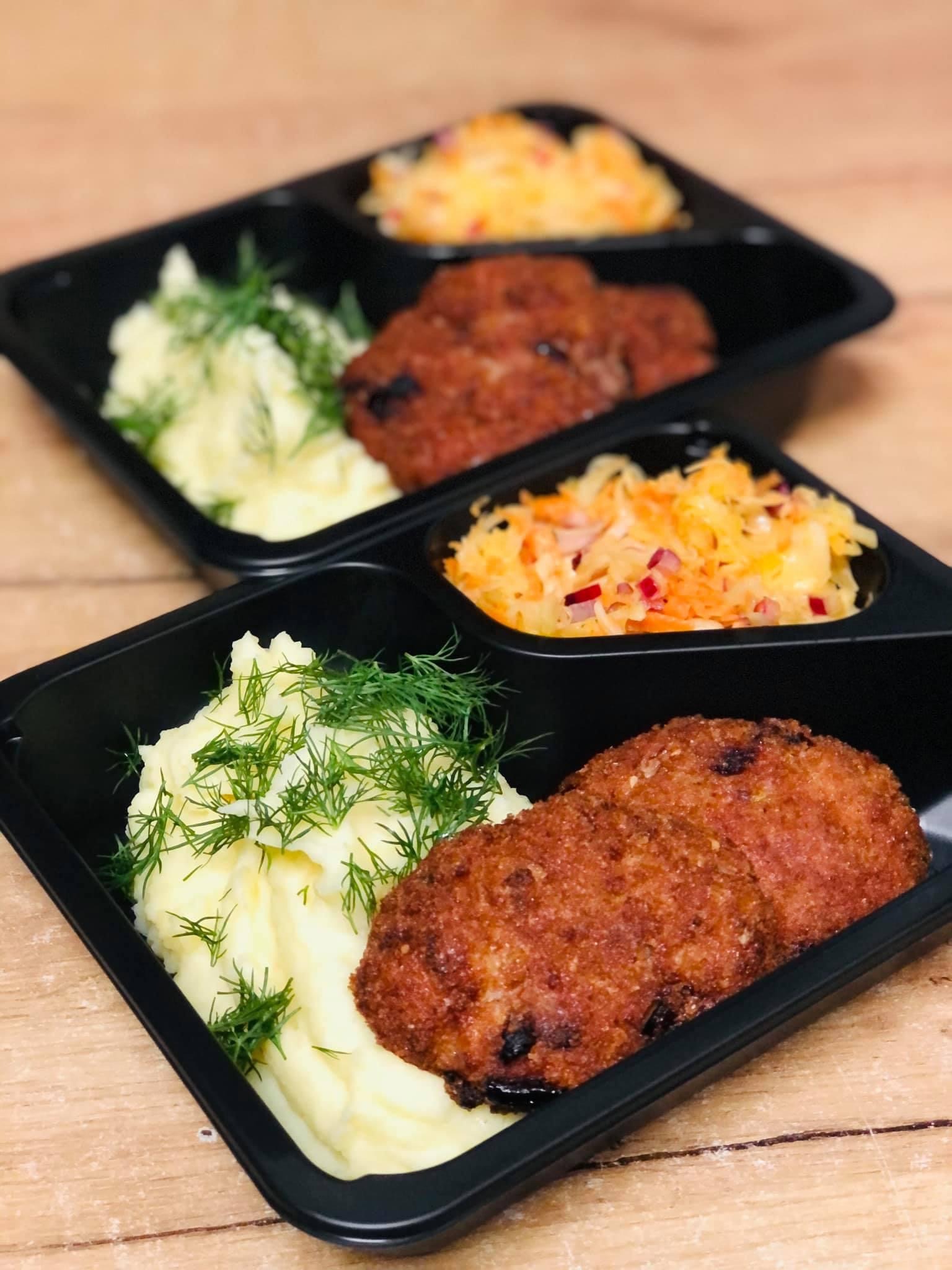 Dwa czarne, trójdzielne pojemniki z jedzeniem: kotlety mielone, ziemniaki puree posypane koperkiem i surówka z marchewki i kapusty z czerwoną cebulą, ułożone na drewnianym stole.