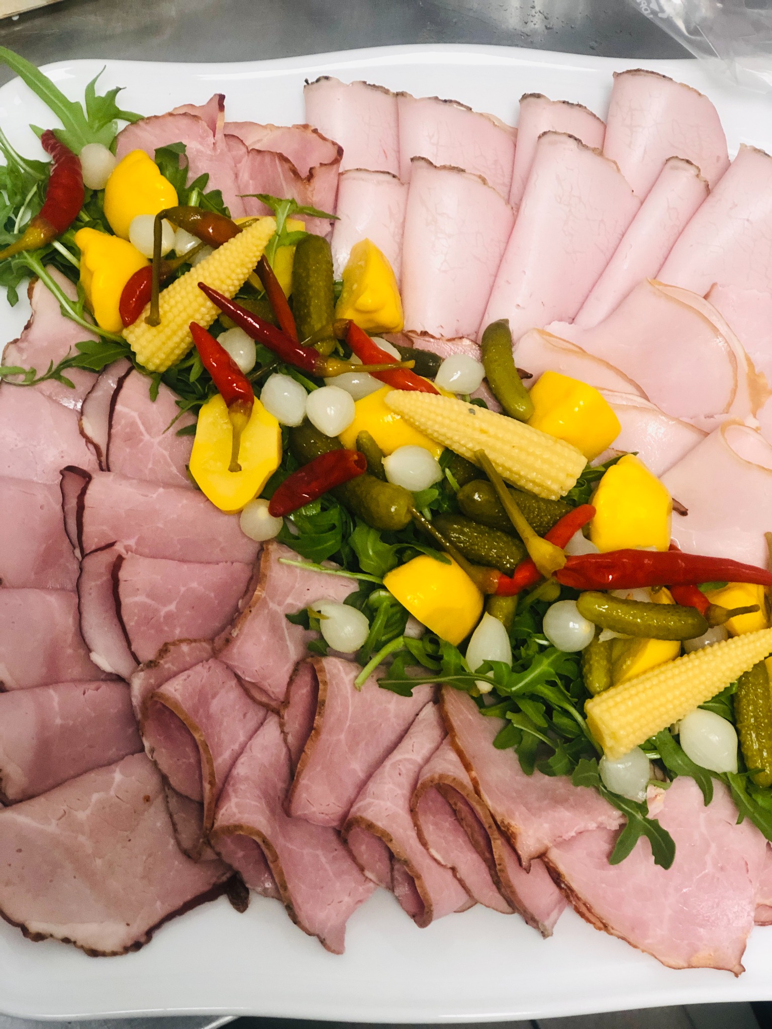 Dekoracyjnie ułożone plastry wędlin z dodatkiem kukurydzy mini, pikli, cebulek i papryczek na białym talerzu.