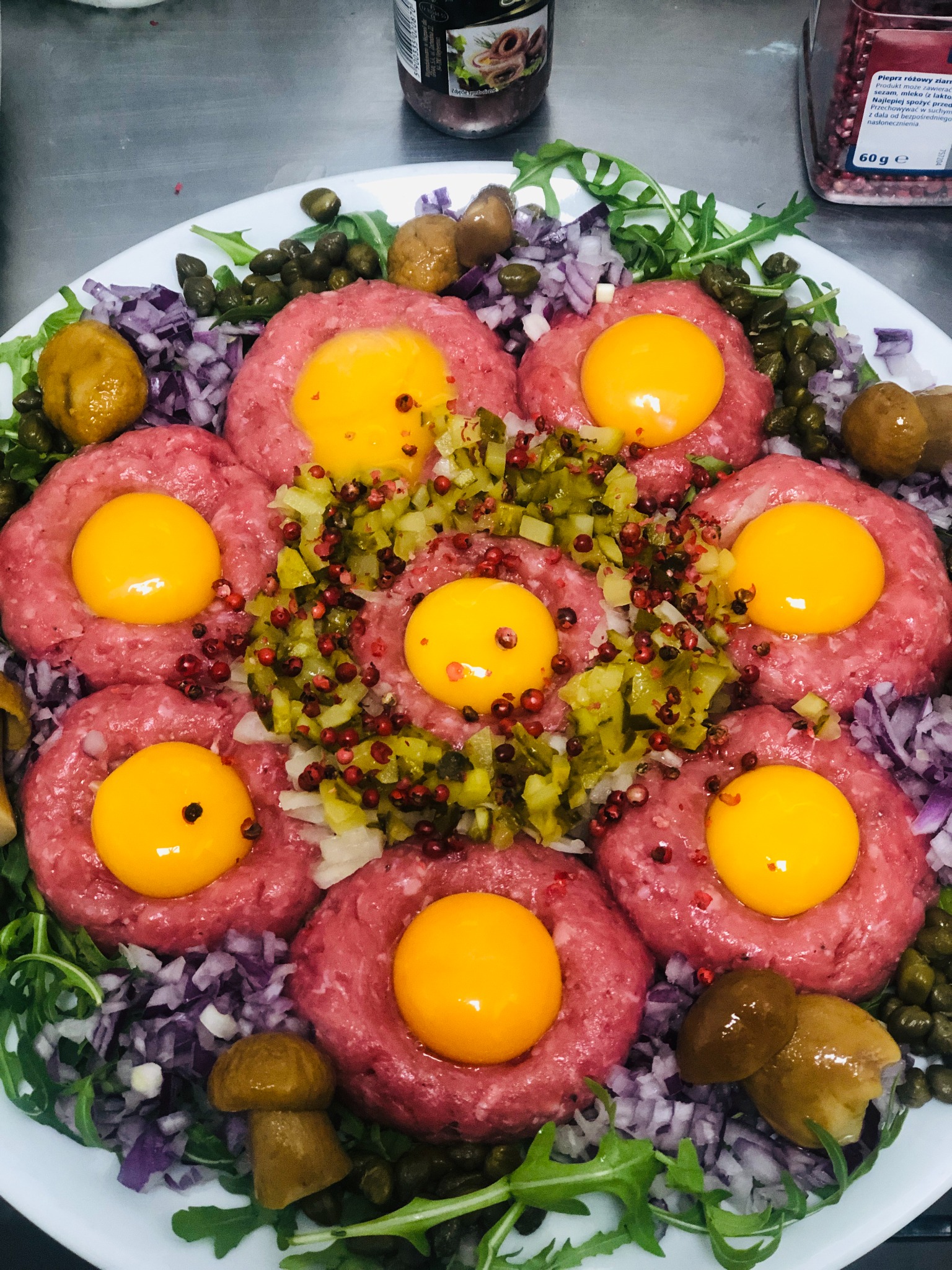 Starannie ułożony na talerzu tatar wołowy z siedmioma porcjami, każda z żółtkiem, otoczony kaparami, marynowanymi grzybami, posiekaną czerwoną cebulą, roszponką i centralnie umieszczoną mieszanką...