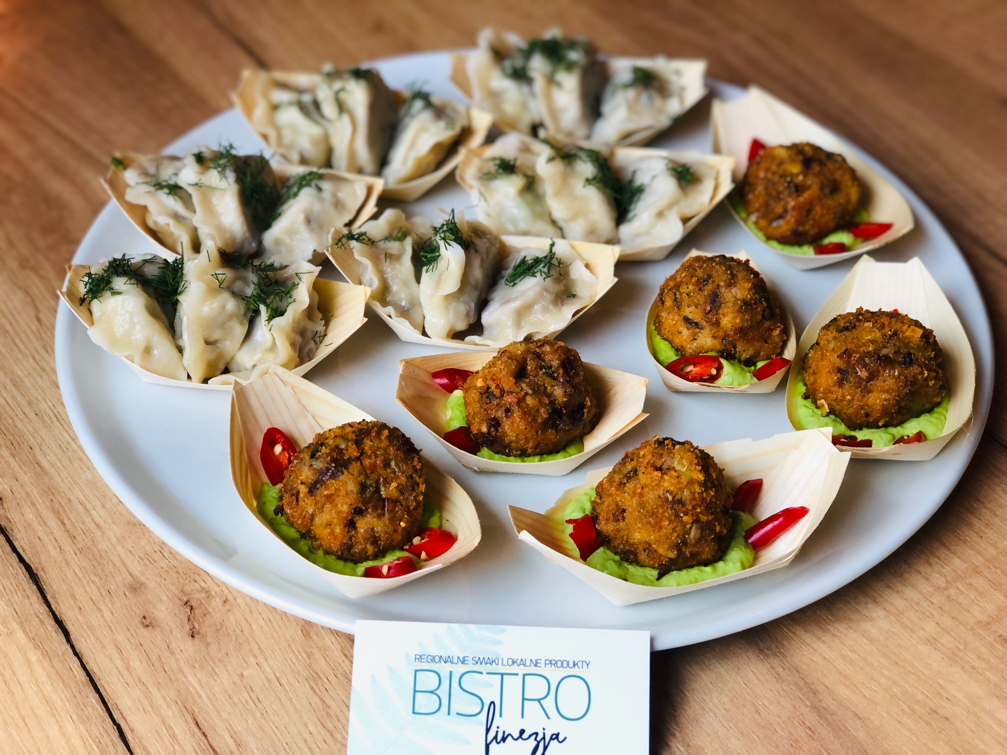 Elegancko podane pierogi z koperkiem i pulpeciki na zielonym puree z papryczką chili, serwowane w drewnianych łódeczkach na białym talerzu, widoczna wizytówka bistro.