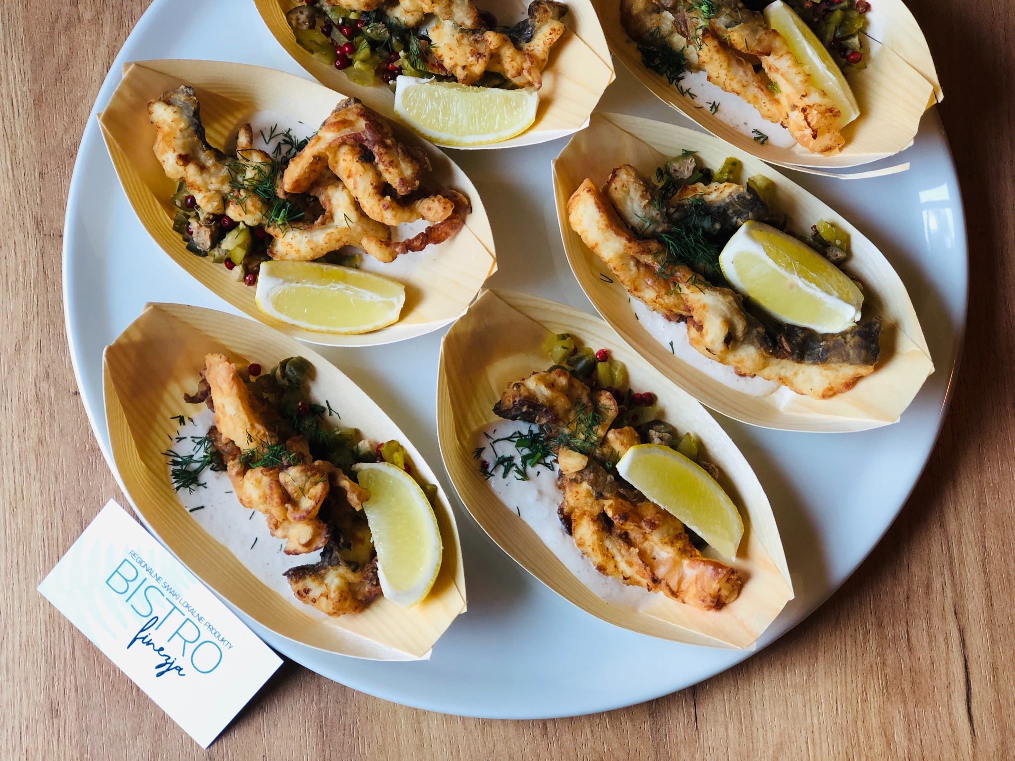 Sześć porcji smażonej ryby w tempurze, podane w drewnianych łódeczkach z sosem, kaparami, czerwoną porzeczką i plasterkiem cytryny na białym talerzu; wizytówka Bistro Finezja w lewym dolnym rogu.