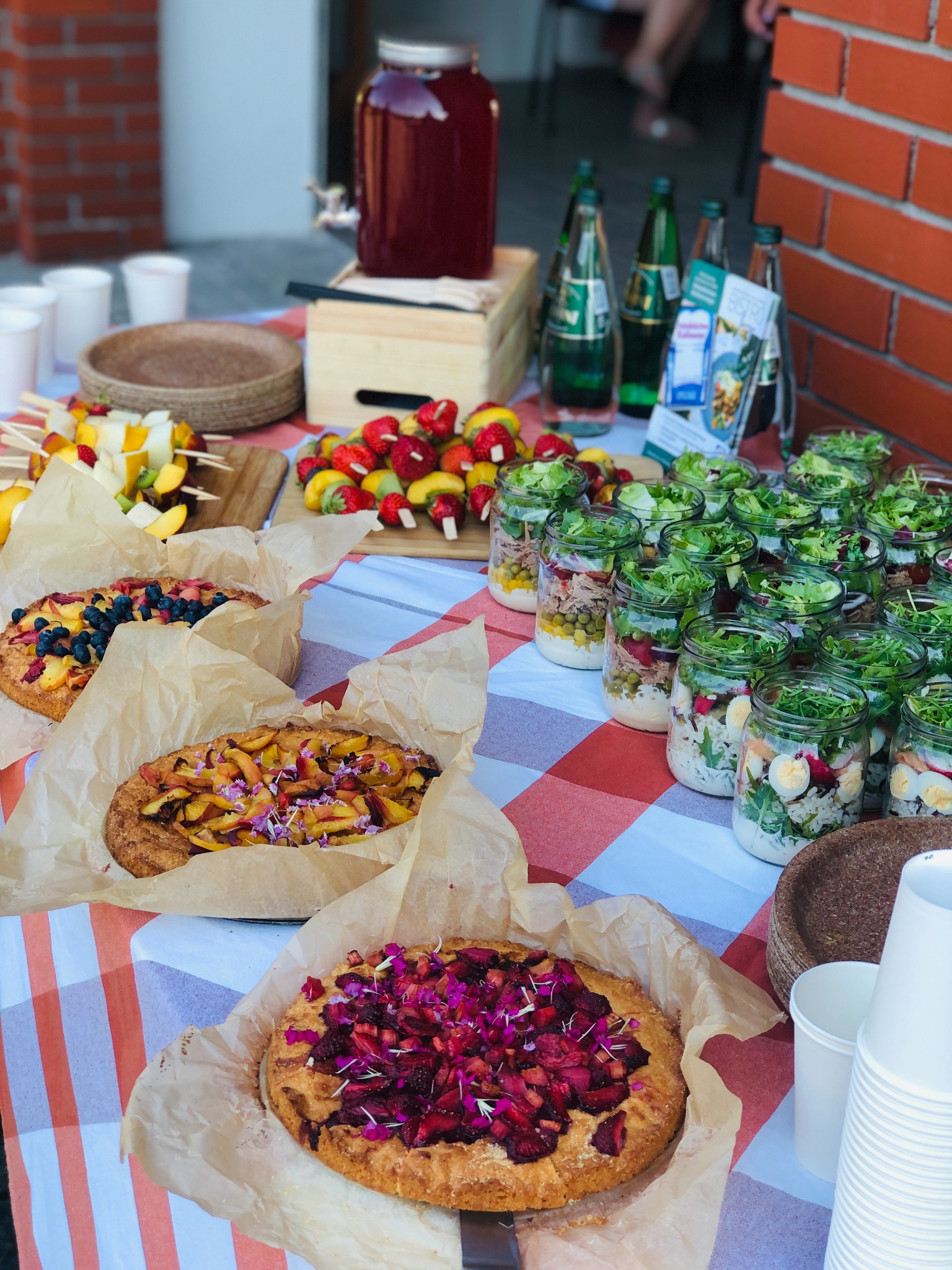 Stół cateringowy z owocowymi szaszłykami, tartami z owocami i sałatkami w słoikach na czerwono-białej kraciastej serwecie.