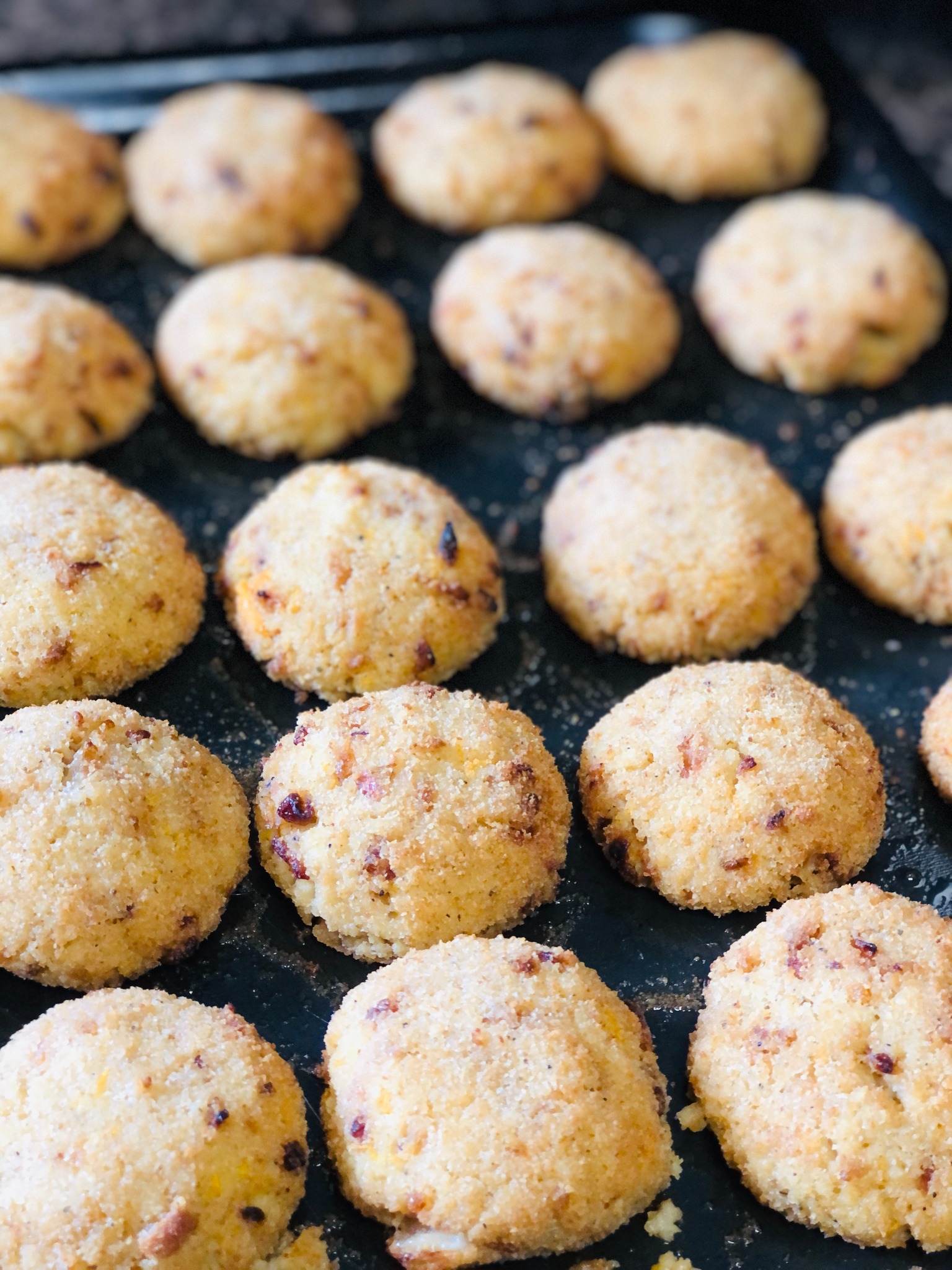 Rządki okrągłych ciasteczek z drobnymi kawałkami owoców na ciemnej blasze do pieczenia, posypane cukrem pudrem.