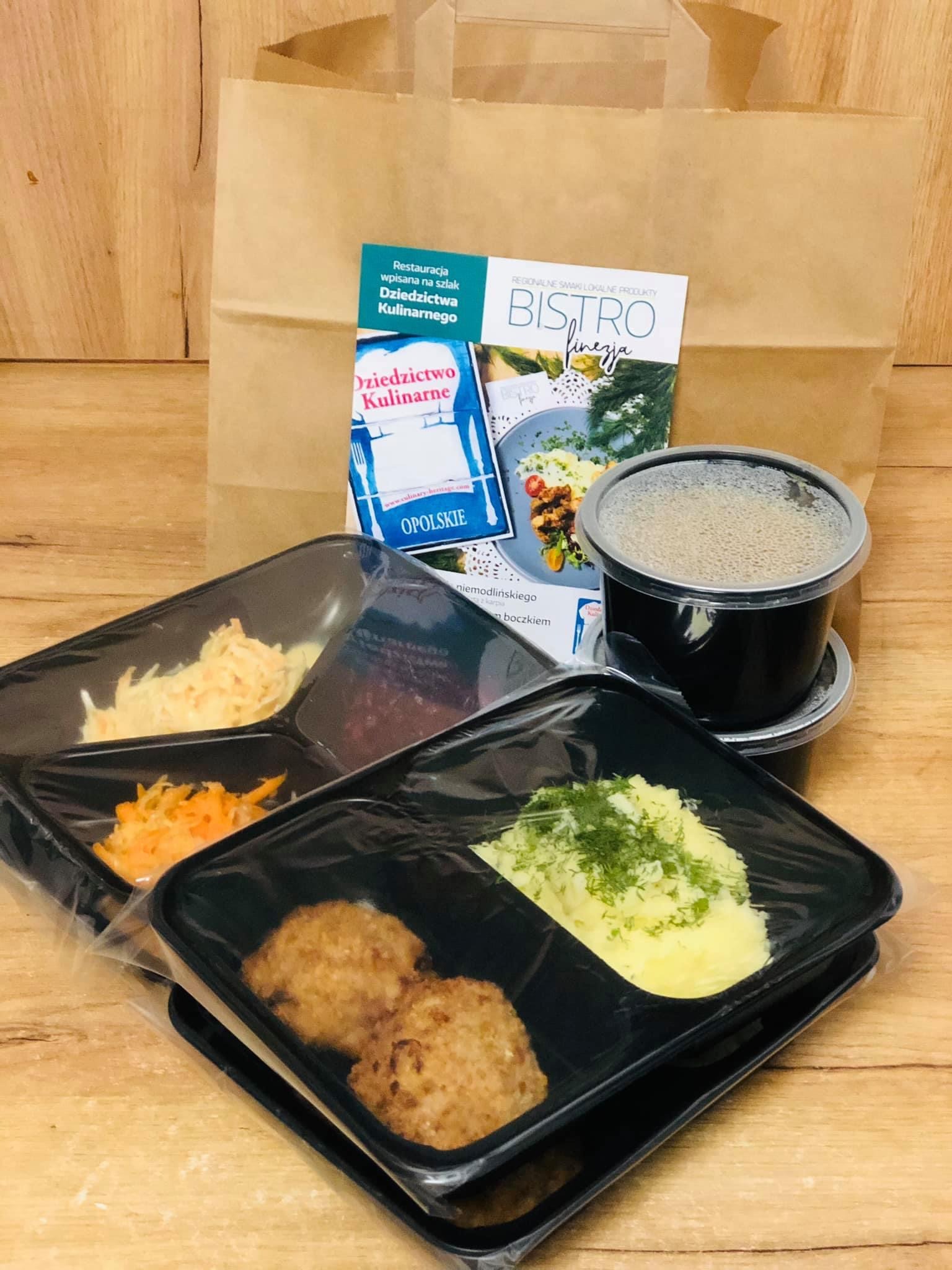 Zestaw obiadowy w czarnych, plastikowych pojemnikach z przezroczystą folią, obejmujący kotlety mielone, ziemniaki z koperkiem, surówki oraz zupę w okrągłym pojemniku, na drewnianym stole, z brązową...