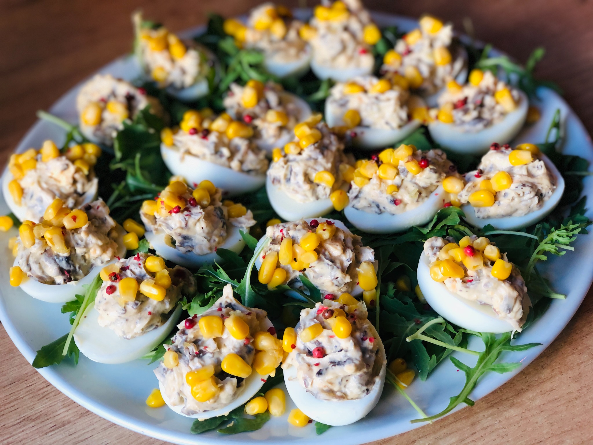 Dekoracyjnie podane jajka faszerowane z farszem z grzybów i kukurydzy, posypane czerwonym pieprzem, ułożone na talerzu z rukolą.