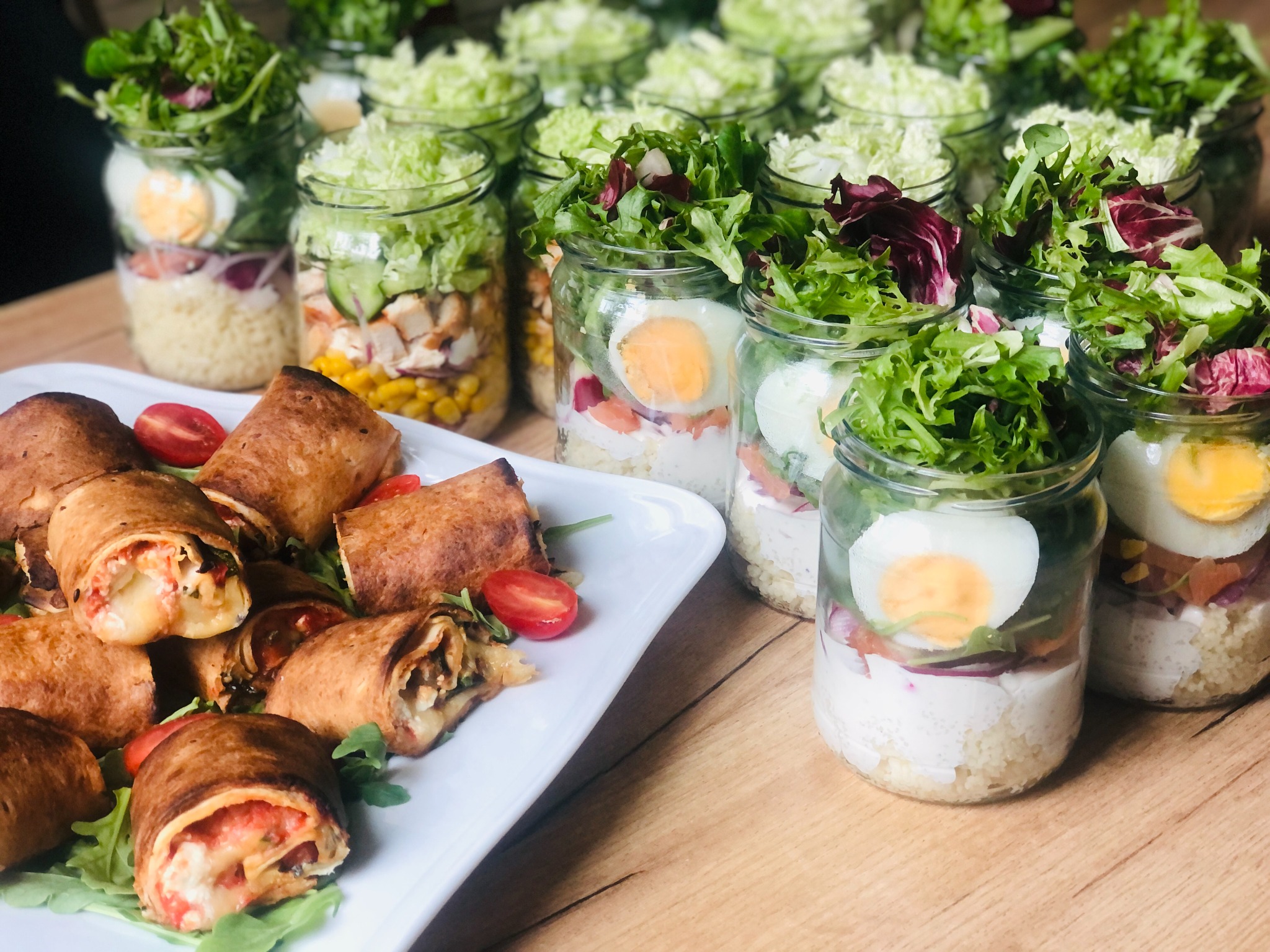 Szereg słoików z warstwowymi sałatkami z jajkiem i zieleniną obok talerza z tortillami pokrojonymi w roladki, udekorowanymi pomidorkami cherry i rukolą.