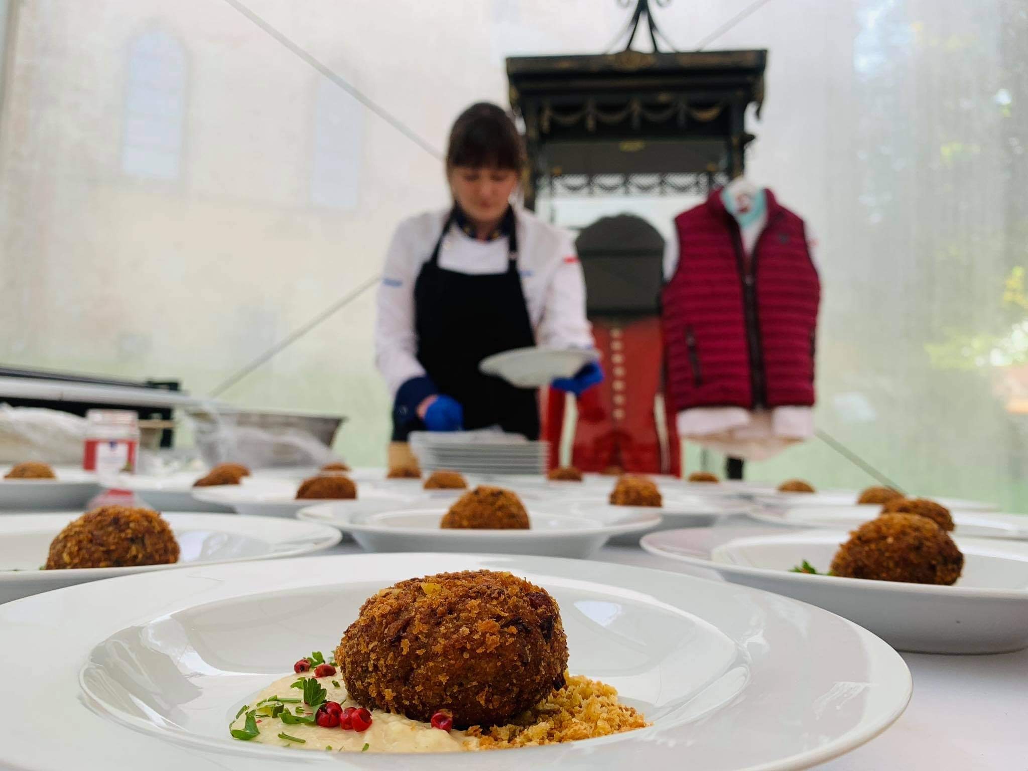 Szef kuchni w białym uniformie i czarnym fartuchu układa na białych talerzach krokiety z sosem i dekoracją z ziół i czerwonych kuleczek, przygotowane do serwowania.
