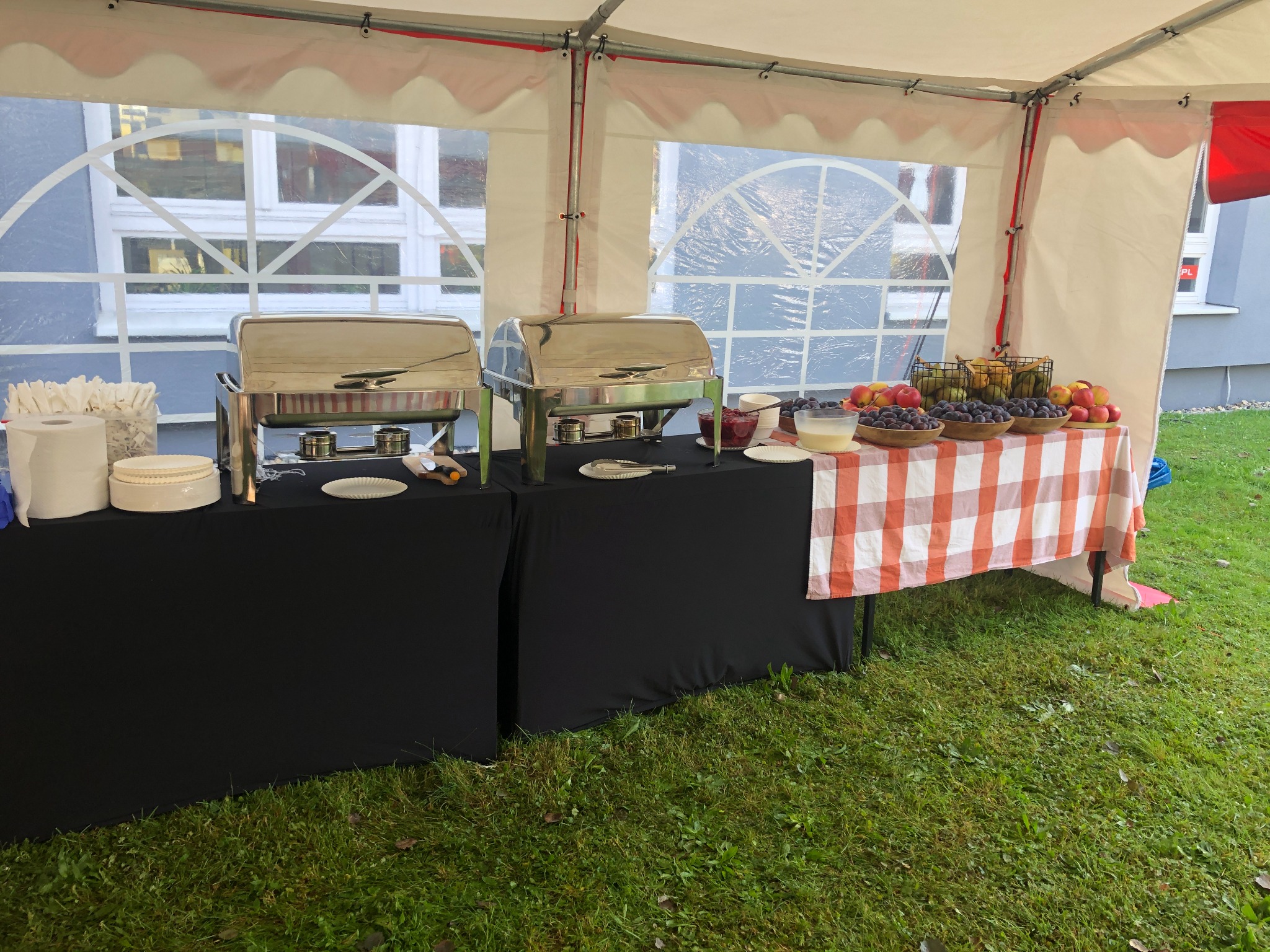 Dwa bufety cateringowe pod namiotem, jeden z podgrzewaczami do potraw, drugi z owocami w drewnianych misach i naczyniami z deserami, na trawniku.