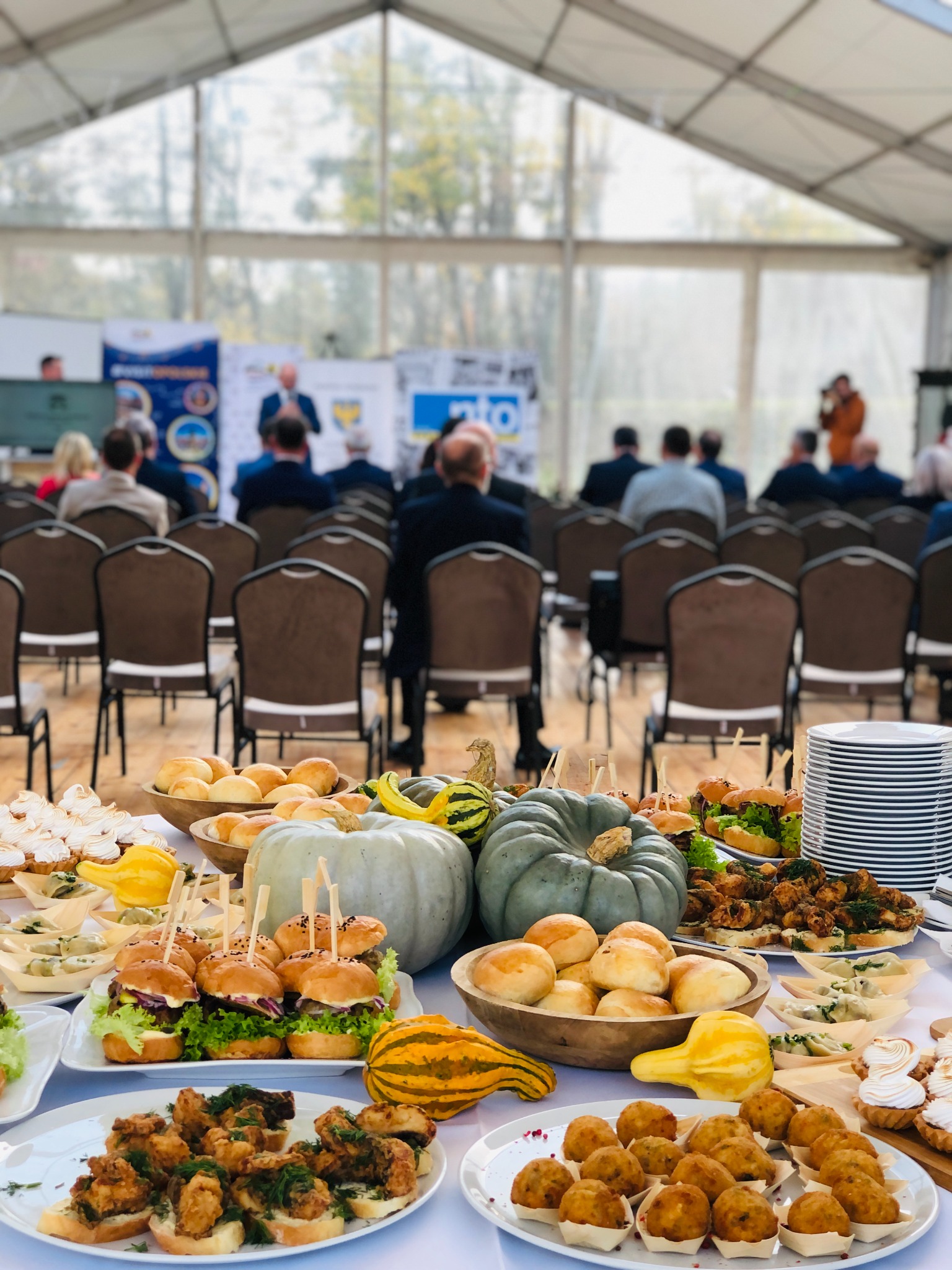 Stół cateringowy z mini burgerami, przekąskami i dyniami dekoracyjnymi na tle sali konferencyjnej z uczestnikami i prelegentami.