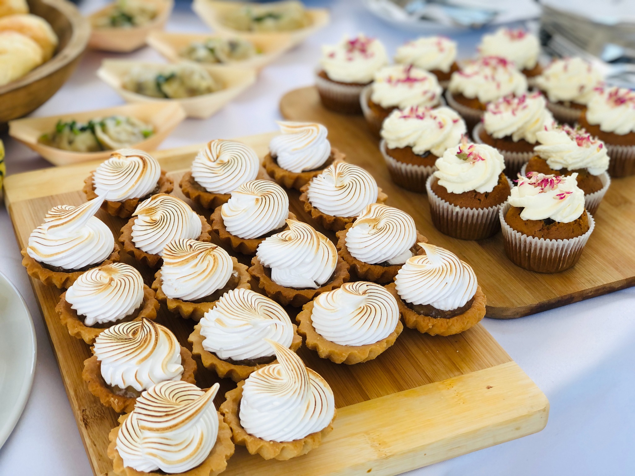 Prezentacja słodkich przekąsek: tartaletki z kremem bezowym z delikatnym przypaleniem i muffinki z białym kremem oraz posypką z płatków róż na drewnianych deskach.