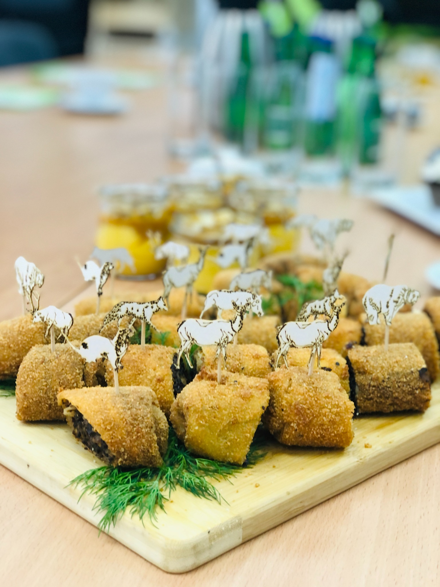 Krokiety z farszem, dekorowane wykałaczkami z motywem zwierząt, ułożone na drewnianej desce z gałązkami koperku, w tle słoiki z marynatami.